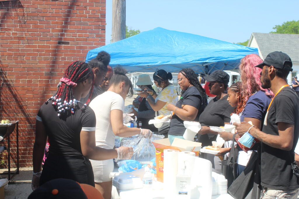 Community event under a blue tent with people serving and receiving food on a bright day